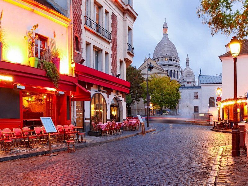 A red-fronted Montmartre café at dusk with the Sacré-Cœur lit behind it